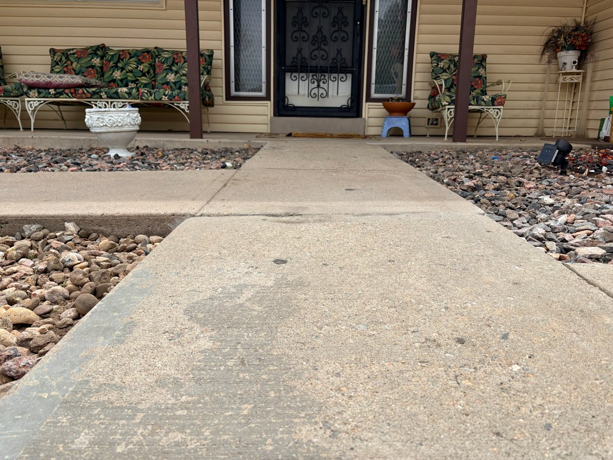 Leveled concrete walkway leading to a front porch with floral cushioned benches, potted plants, and a black iron security door.