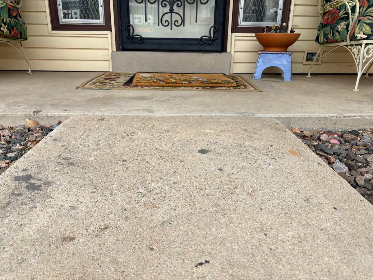 Repaired concrete walkway leading to a porch with doormats, a black door, and cushioned chairs.