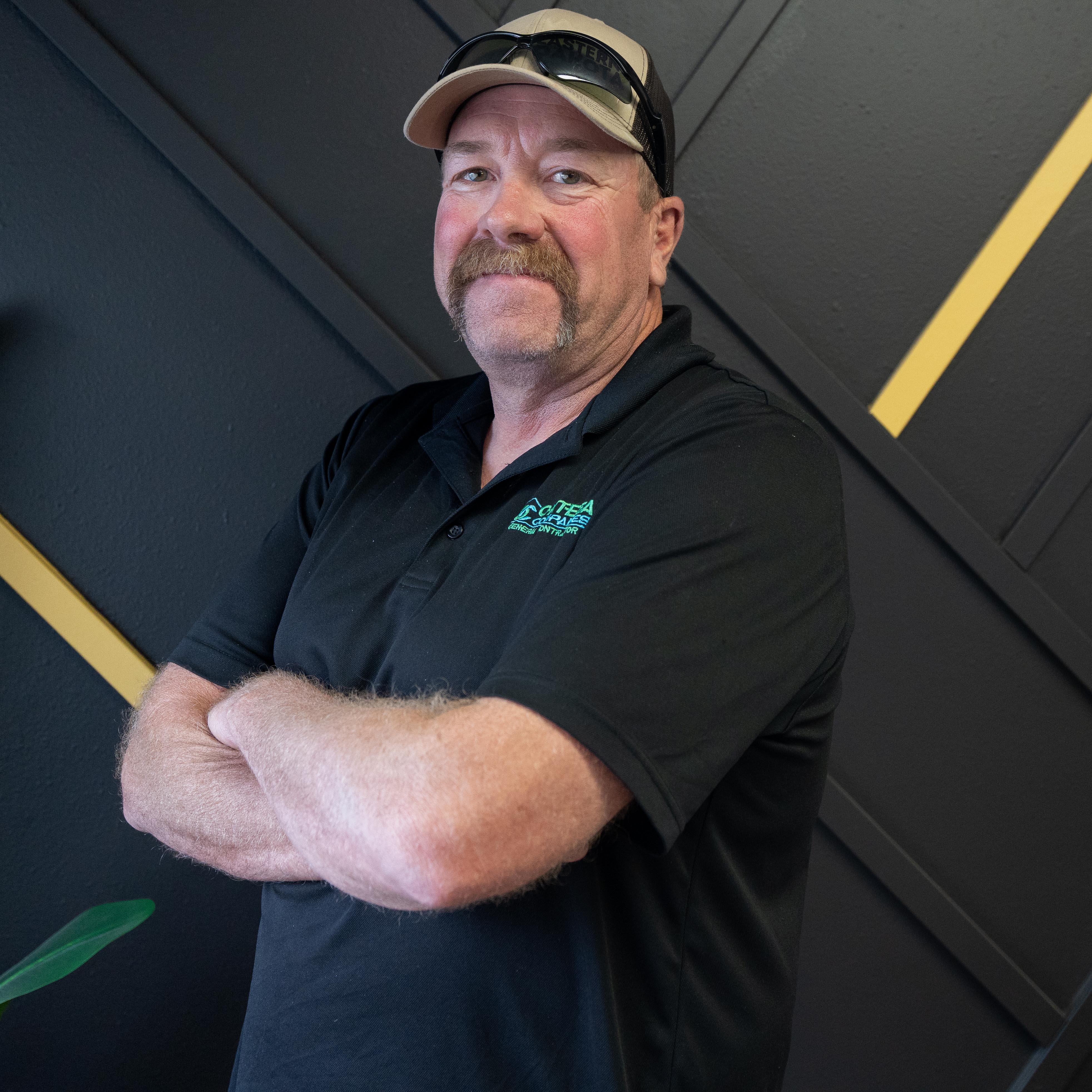 Man with a mustache and cap crossing his arms, standing in front of a dark wall with diagonal yellow stripes.