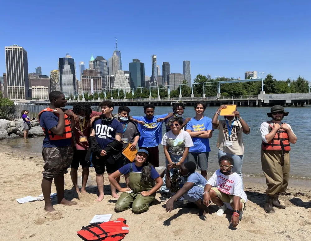 Teen students and instructors pose on a small sandy cove after field work, the Lower Manhattan skyline behind
