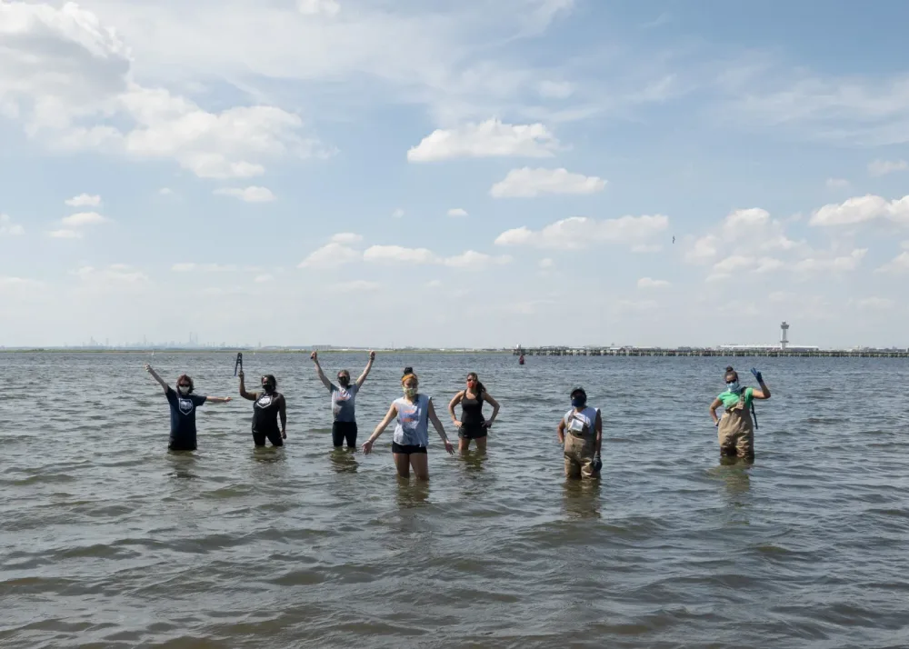 Seven masked volunteers stand waist-deep in bay water with arms raised, an airport control tower on the horizon