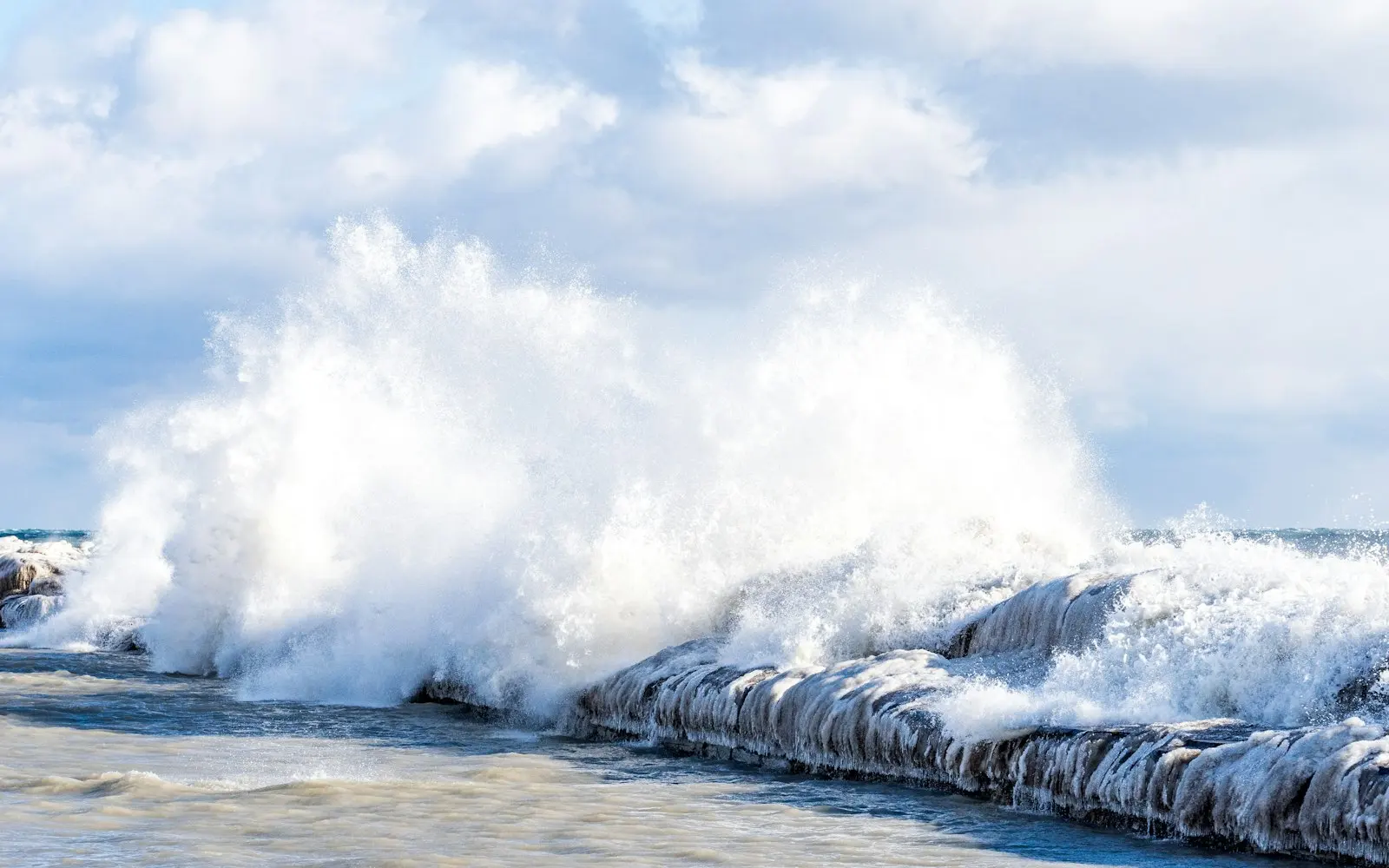 Waves crash over an ice-covered breakwater under a cloudy sky