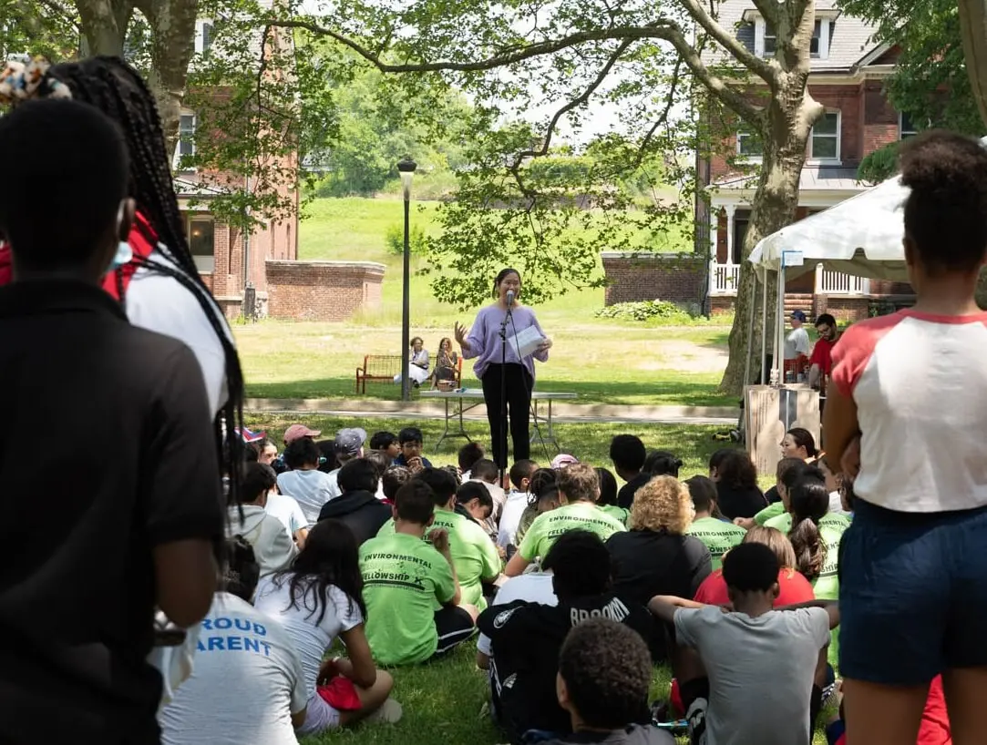 A speaker addresses students seated on a lawn, many in green Environmental Fellowship shirts