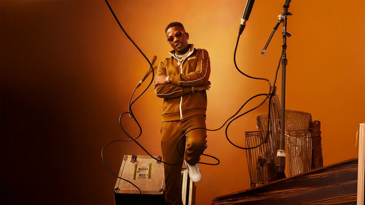 Person in a mustard-colored jacket, standing confidently with arms crossed in front of an orange background, framed by wires or cables.