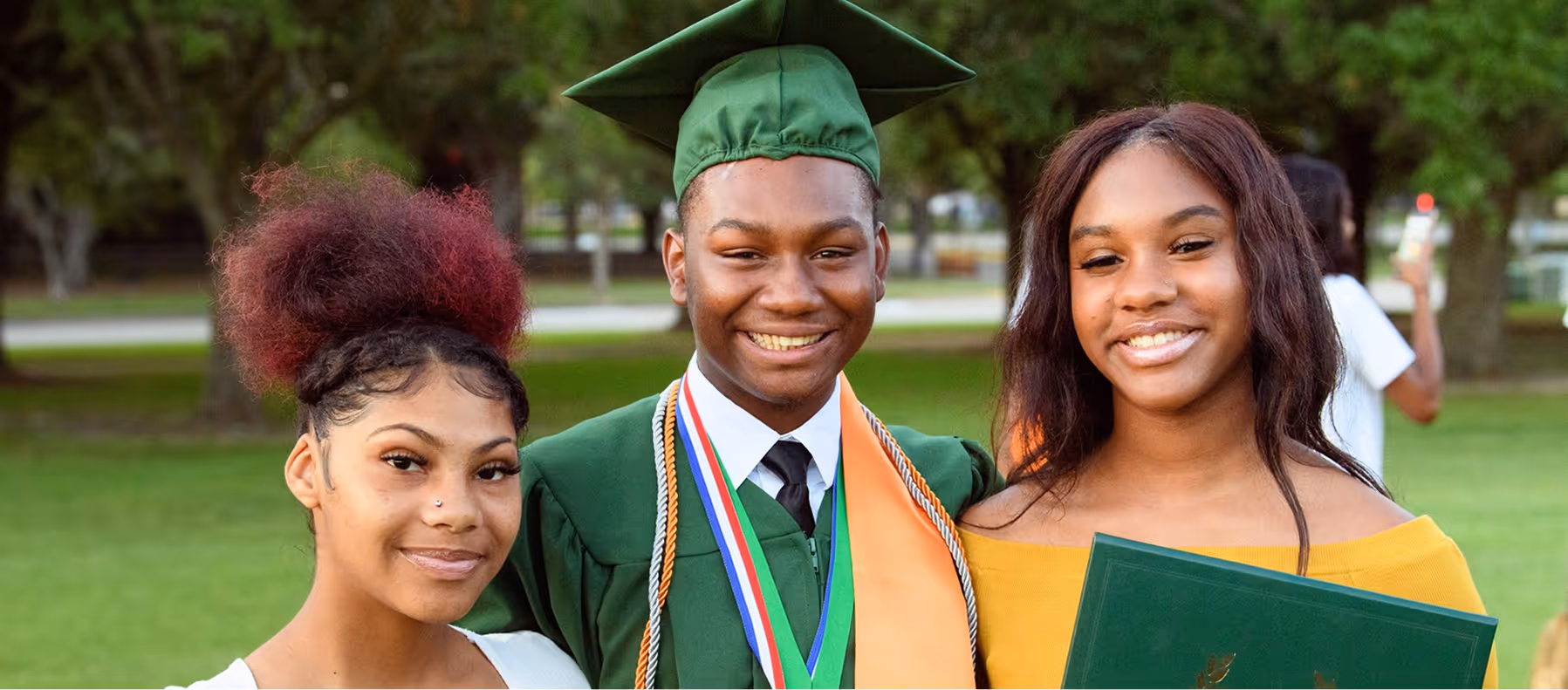 Three smiling young adults standing outdoors, one wearing a green graduation cap and gown with medals.
