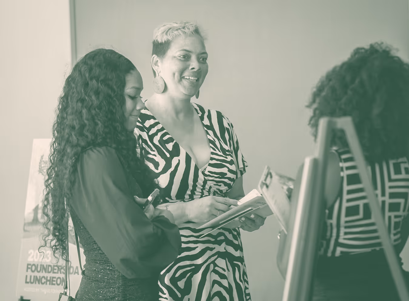 Three women engaged in conversation, one holding a notebook, at an indoor event with a '2023 Founders Day Luncheon' sign in the background.