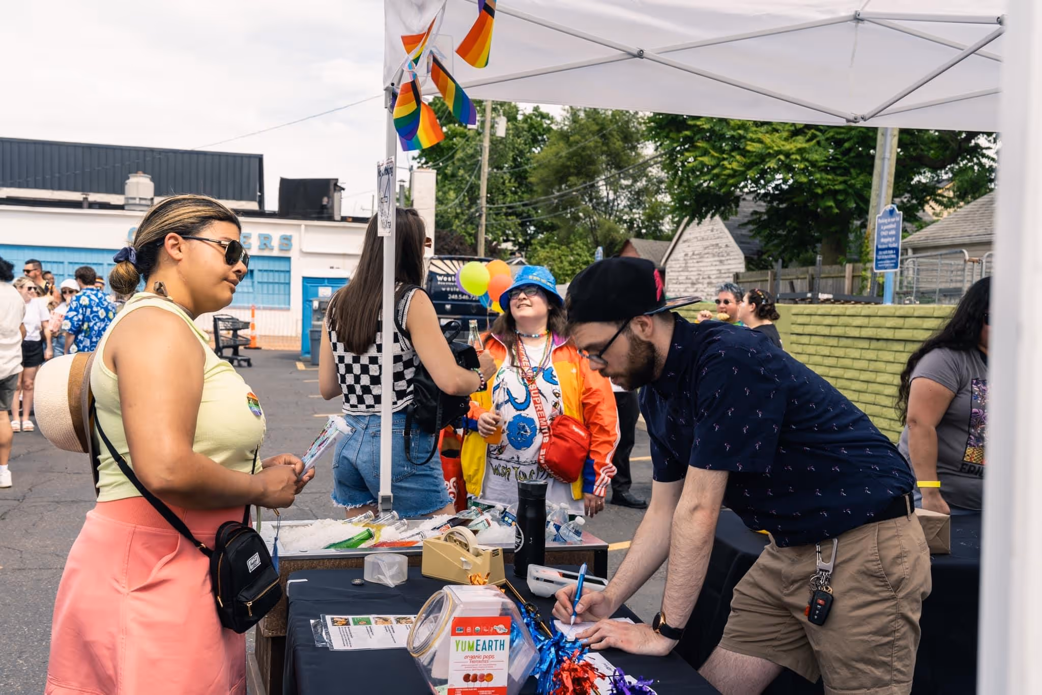 A guest signs up at a vendor table while others stand nearby under a tent decorated with rainbow flags and balloons.