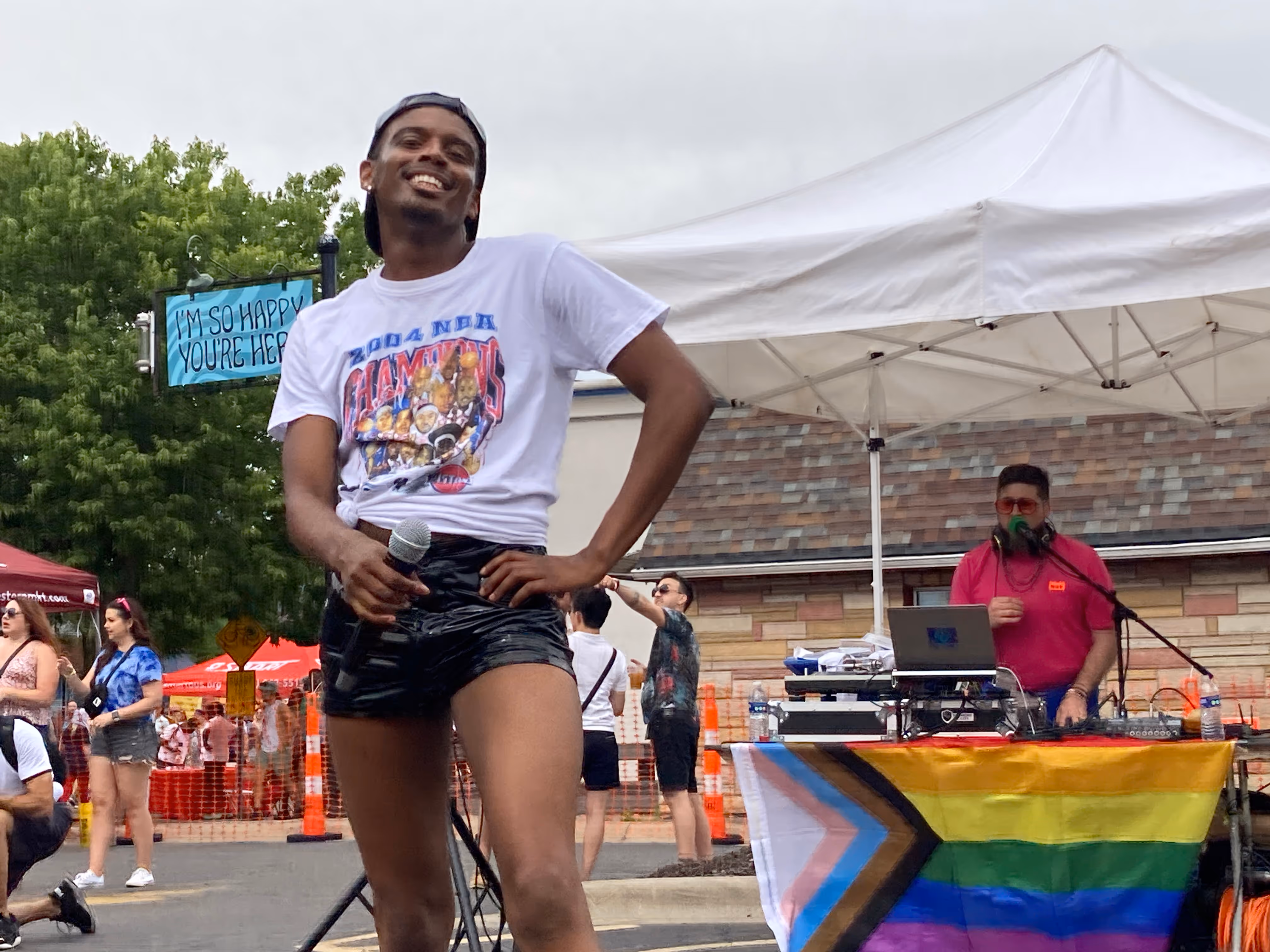 A performer smiles and holds a microphone in front of a DJ booth decorated with a Pride flag, entertaining the crowd.