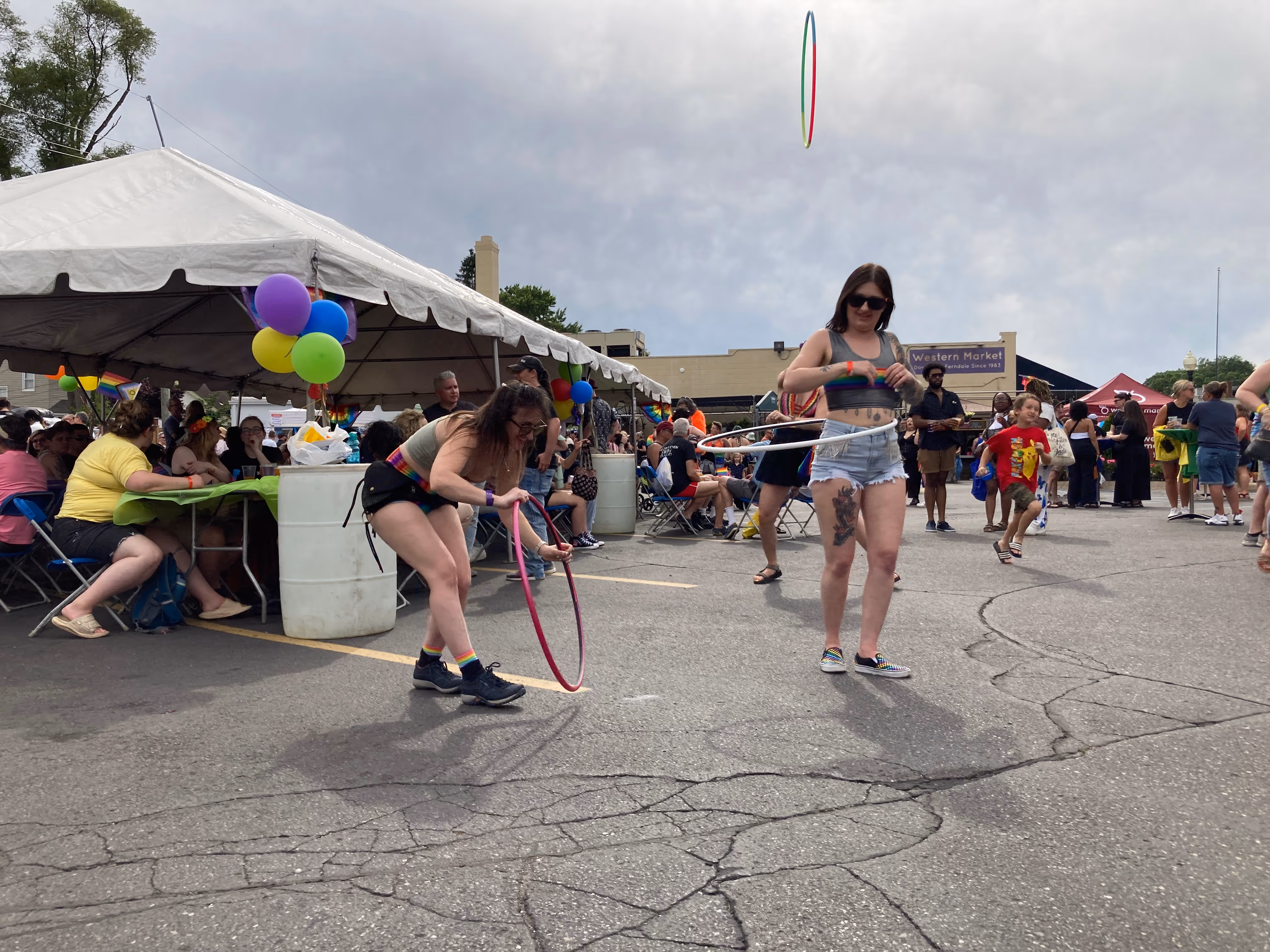 A hula hoop toss is midair as participants laugh and play a game, surrounded by a lively Pride crowd and colorful decorations.