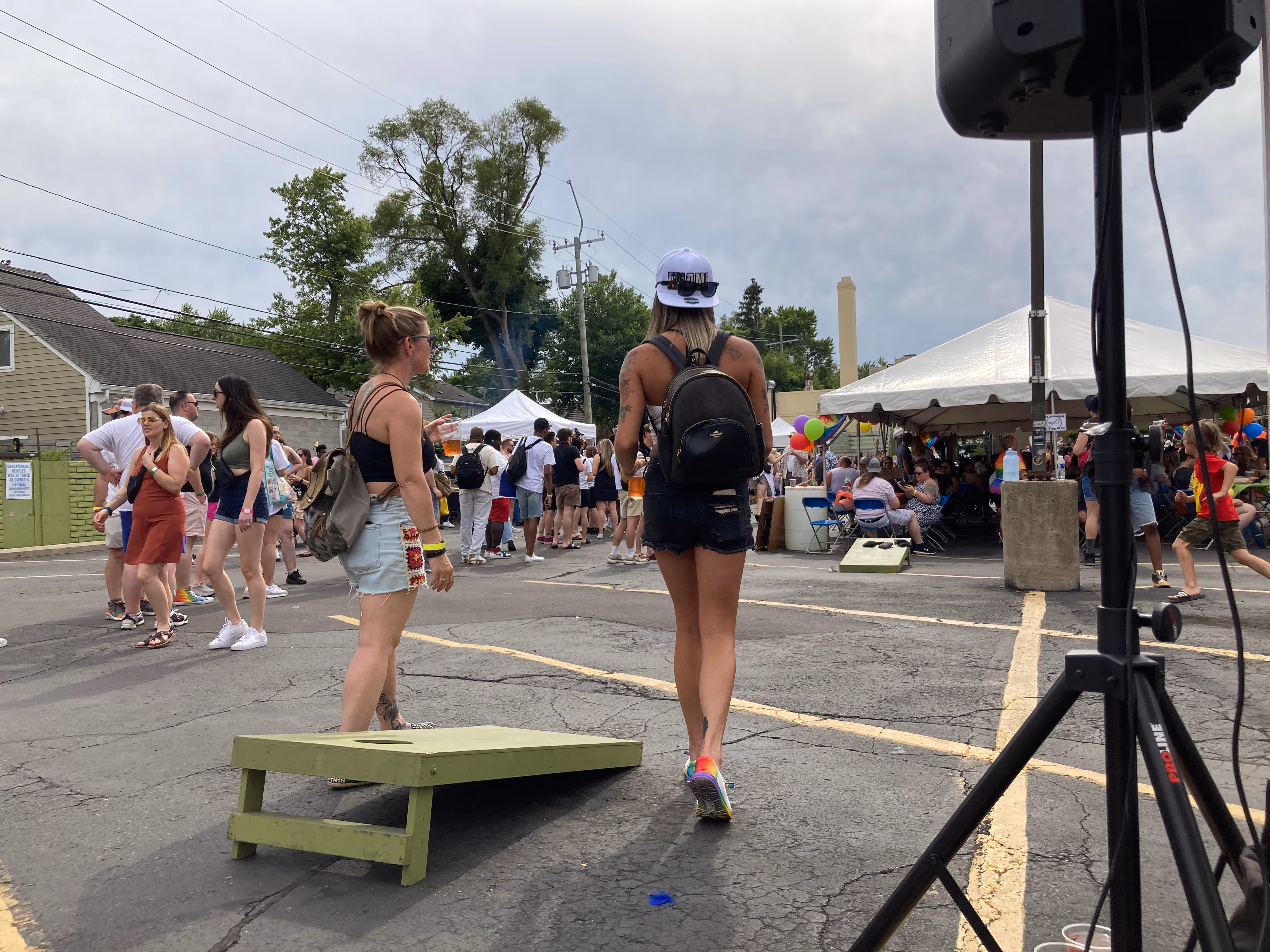 Two people holding drinks walk toward a busy tent area while others mingle and play games in the Western Market parking lot during Pride.