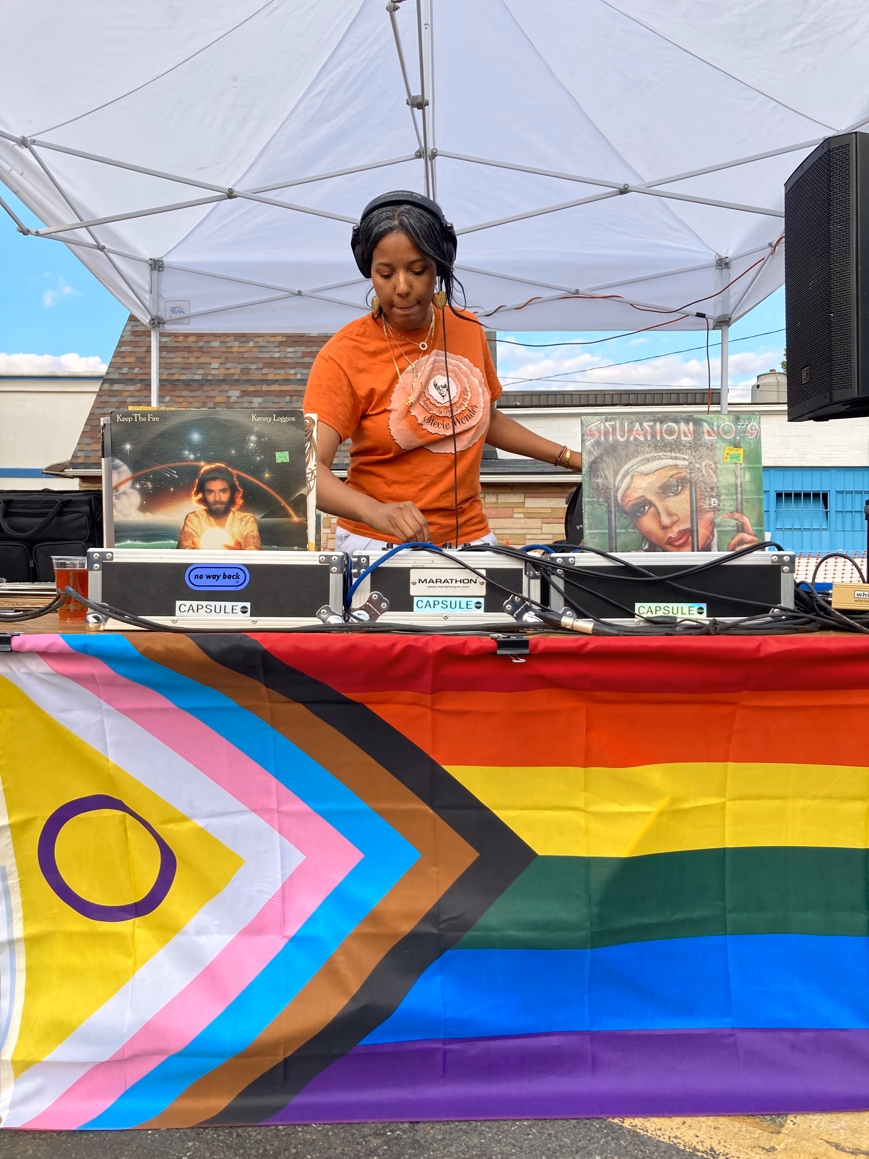 A DJ stands behind turntables draped in a Pride flag, smiling and playing music under a tent at the outdoor event.