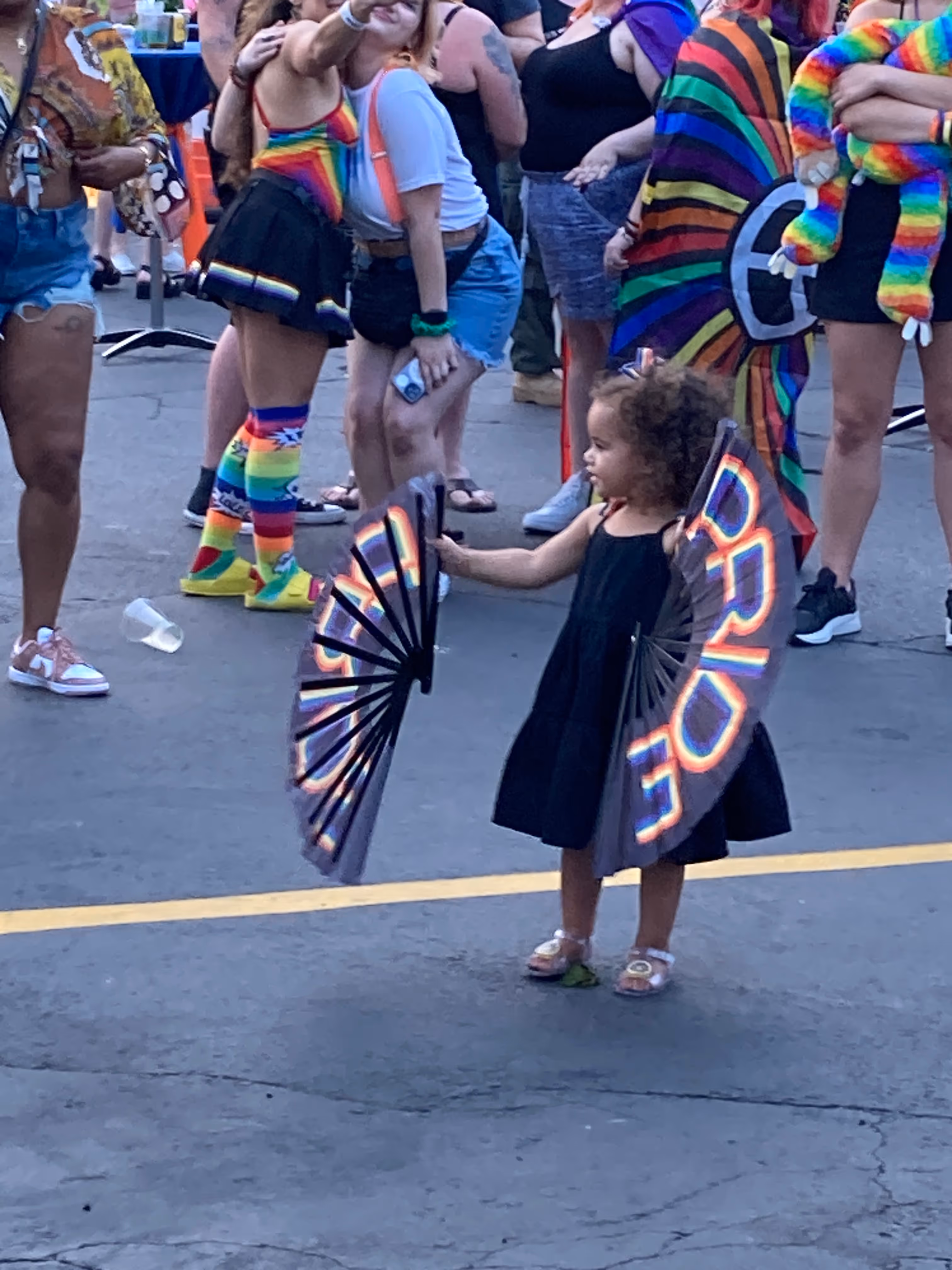 A young child in a black dress stands in the middle of a Pride celebration holding two large rainbow “PRIDE” fans, while a colorful crowd in festive outfits gathers and takes photos behind her.