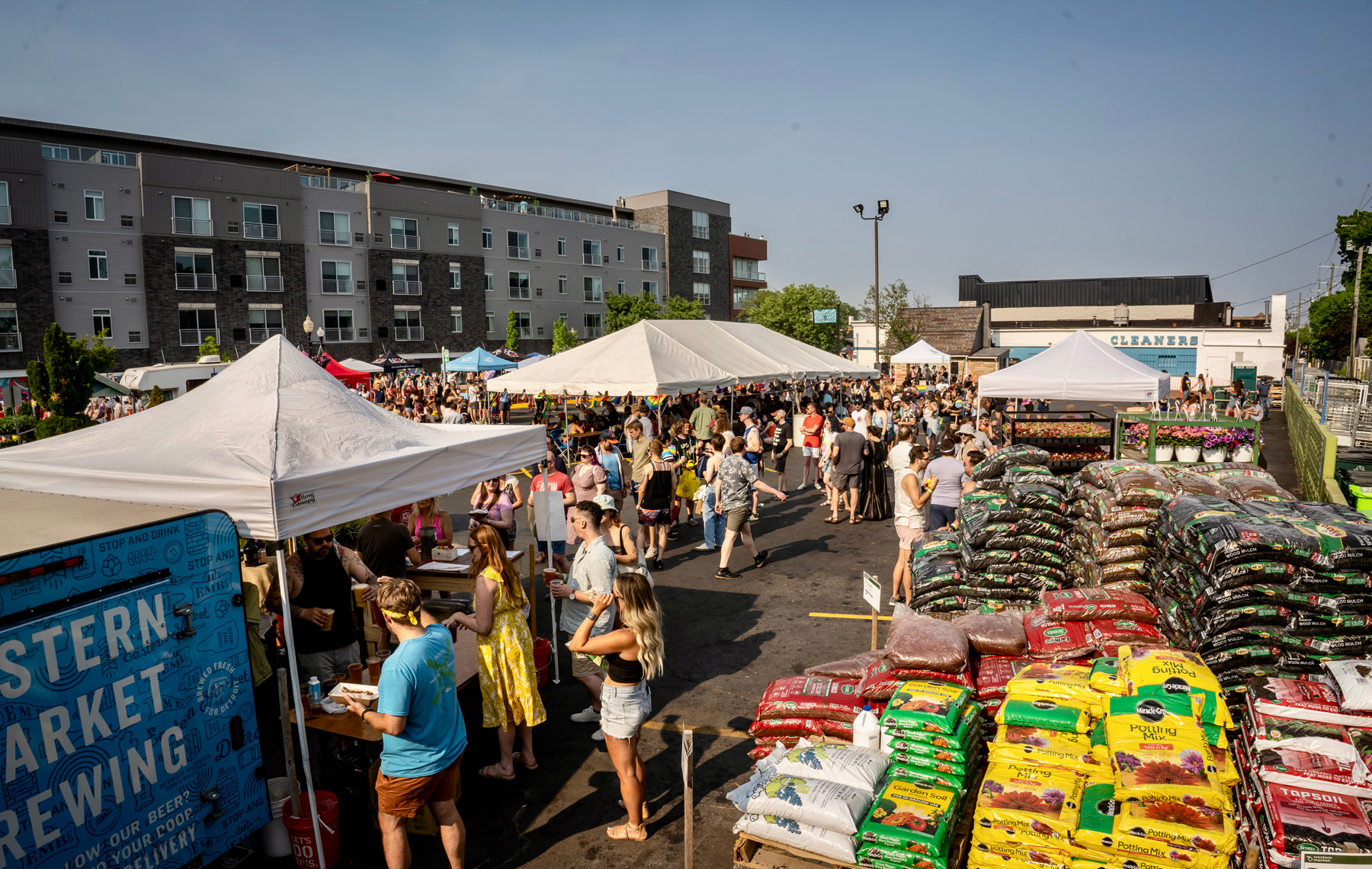 A large crowd gathers under white tents in Western Market’s parking lot during a sunny Pride celebration, with vendors, drinks, and community members filling the space.