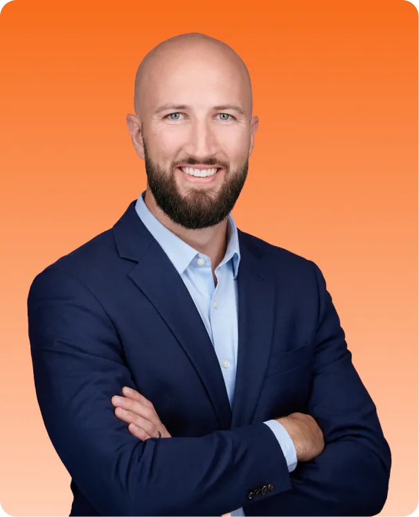 Smiling bald man with beard wearing a navy blazer and light blue shirt standing with arms crossed against an orange background.