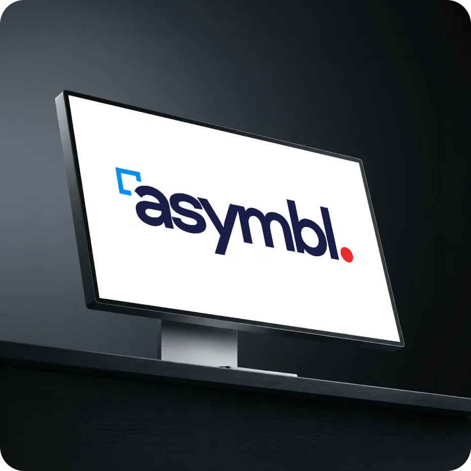 Computer monitor on a dark desk displaying the word 'asymbl.' with a blue square and red dot.