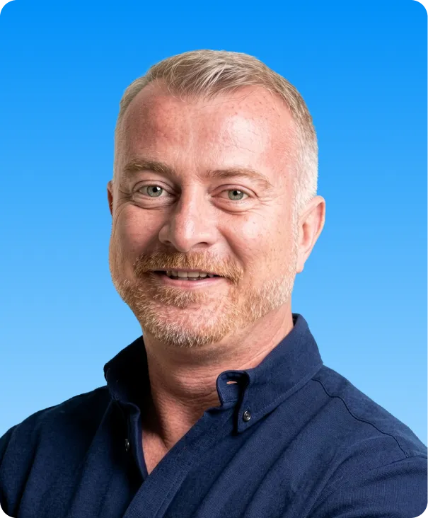 Smiling middle-aged man with short light hair and beard wearing a dark blue collared shirt against a blue gradient background.