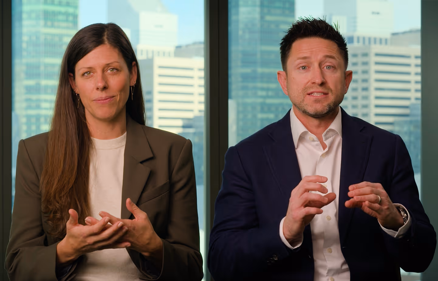 A woman in a brown blazer and white sweater and a man in a dark blazer and white shirt sit side by side with city buildings visible through windows behind them, both gesturing with their hands.