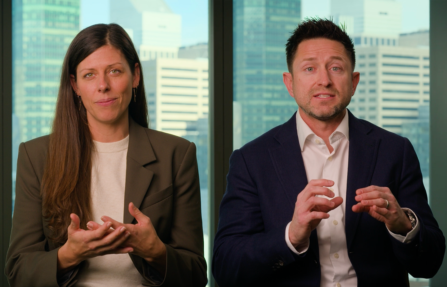 A woman in a brown blazer and white sweater and a man in a dark blazer and white shirt sit side by side with city buildings visible through windows behind them, both gesturing with their hands.