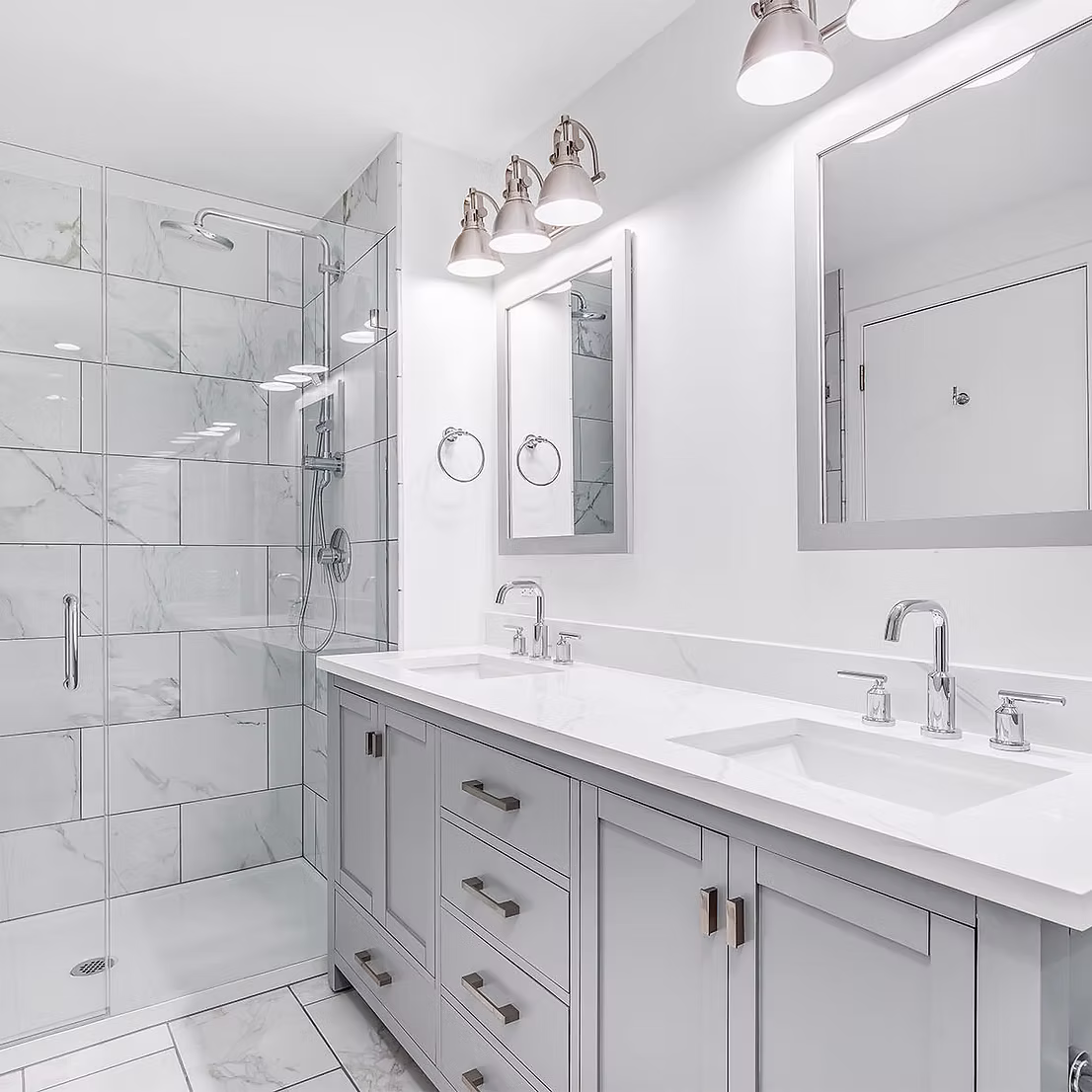 Luxury master bathroom remodel featuring a gray double vanity with white quartz countertops, chrome fixtures, and a walk-in glass shower. The shower and floor are finished with large-format white marble tiles, complemented by industrial-style vanity lighting and framed mirrors.