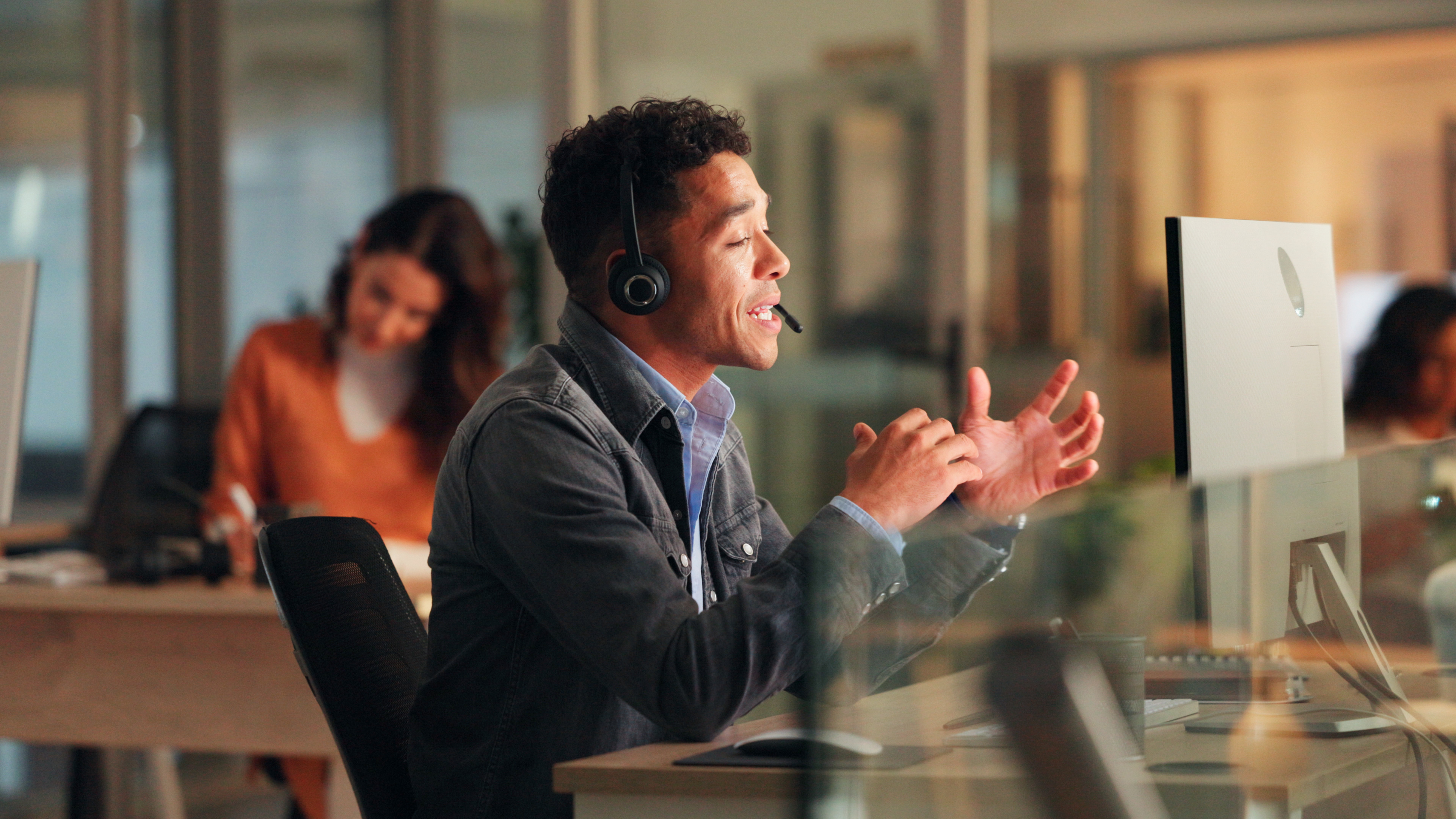 Young man wearing a headset speaking and gesturing at a computer in a modern office with colleagues working in the background.