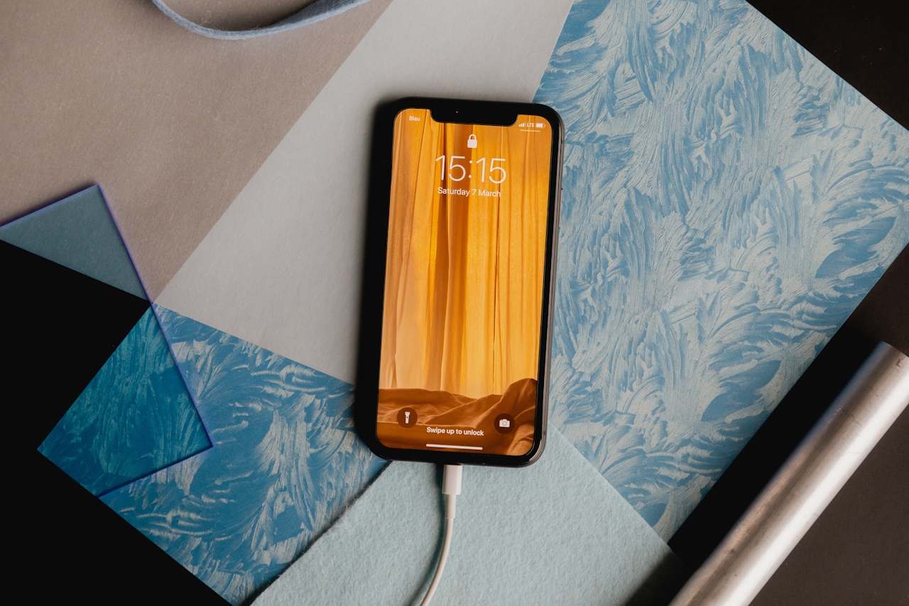 Smartphone displaying locked screen with time 15:15 and date Saturday 7 March, lying on textured blue and beige papers with a charging cable connected.