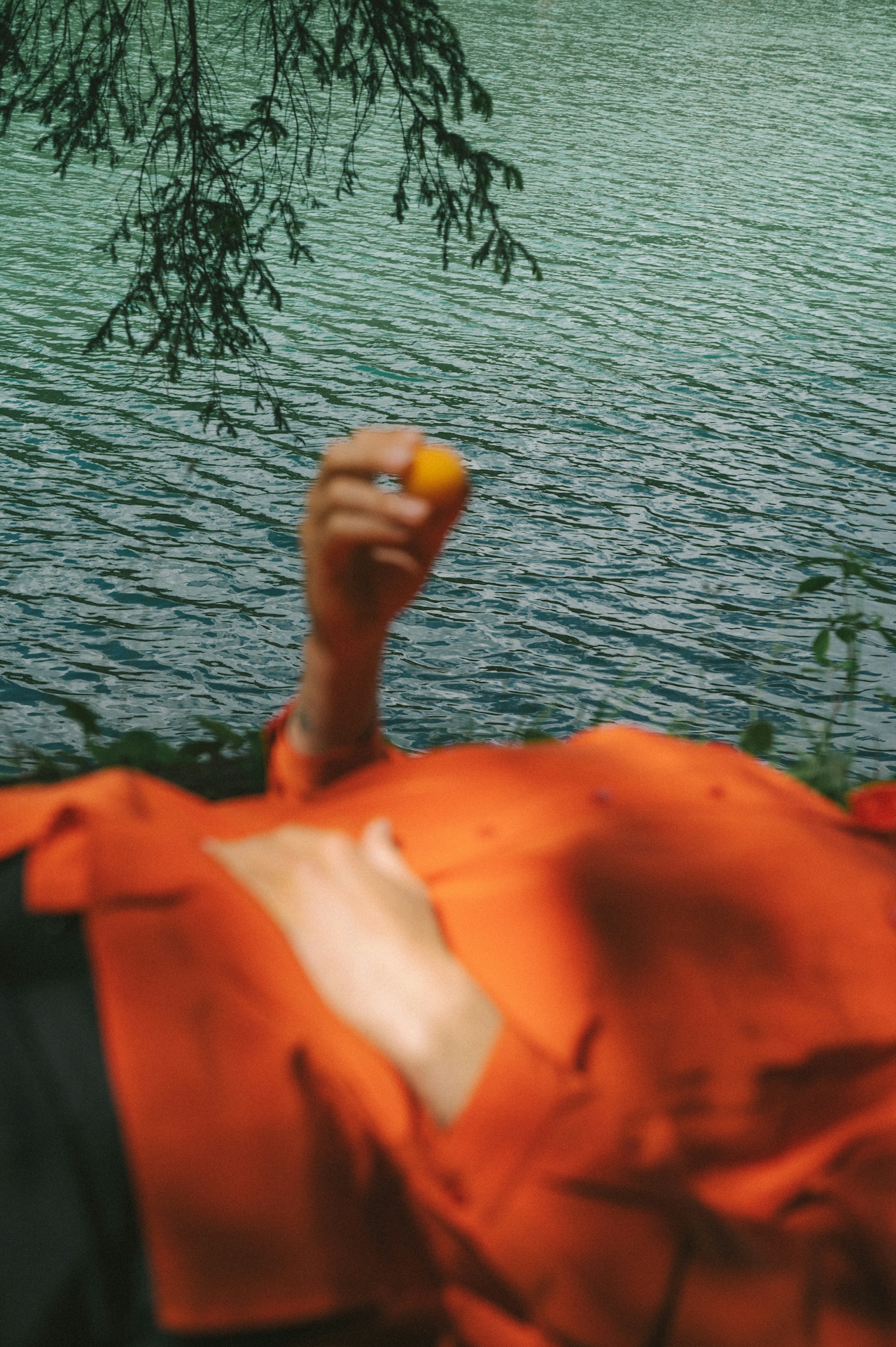 Person lying down near water with tree branches overhead, holding a small orange fruit.