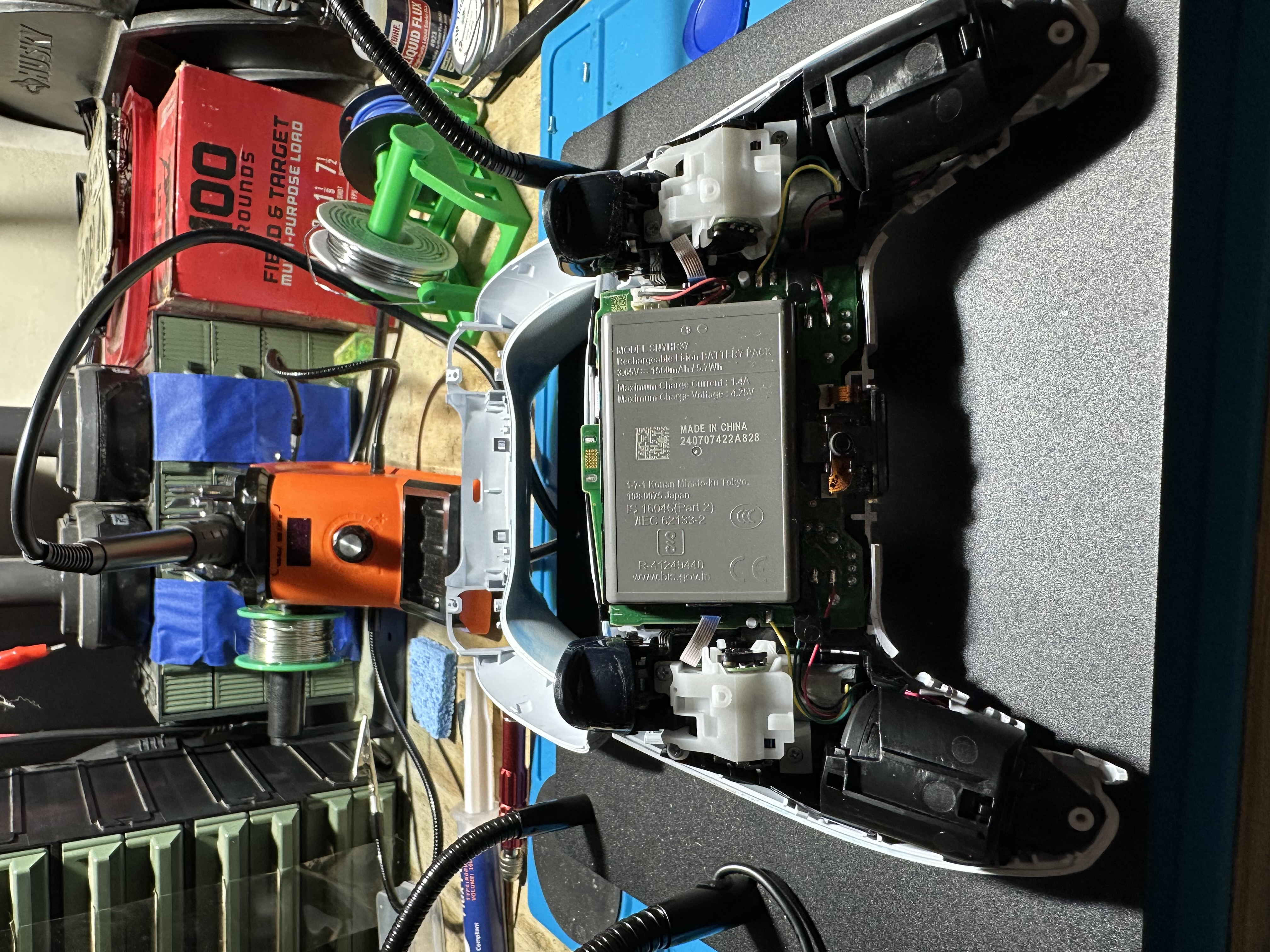 Opened white game controller on a workbench with circuit board and battery exposed, surrounded by soldering tools and supplies.