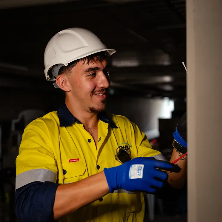 Chill Merchants apprentice in hi-vis, hard hat, and blue gloves smiling while working on electrical connections in a commercial plant room