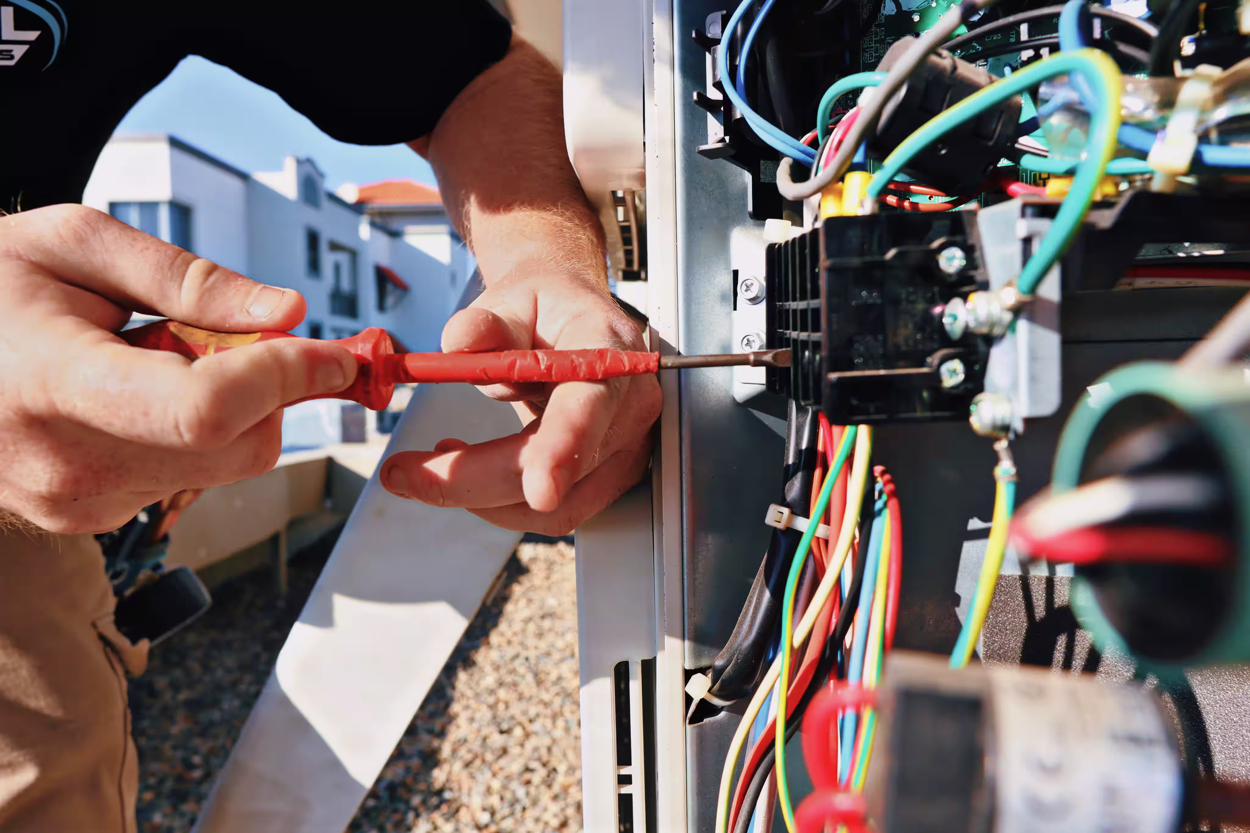 Technician using a red screwdriver to work on colorful wiring inside an electrical panel outdoors.