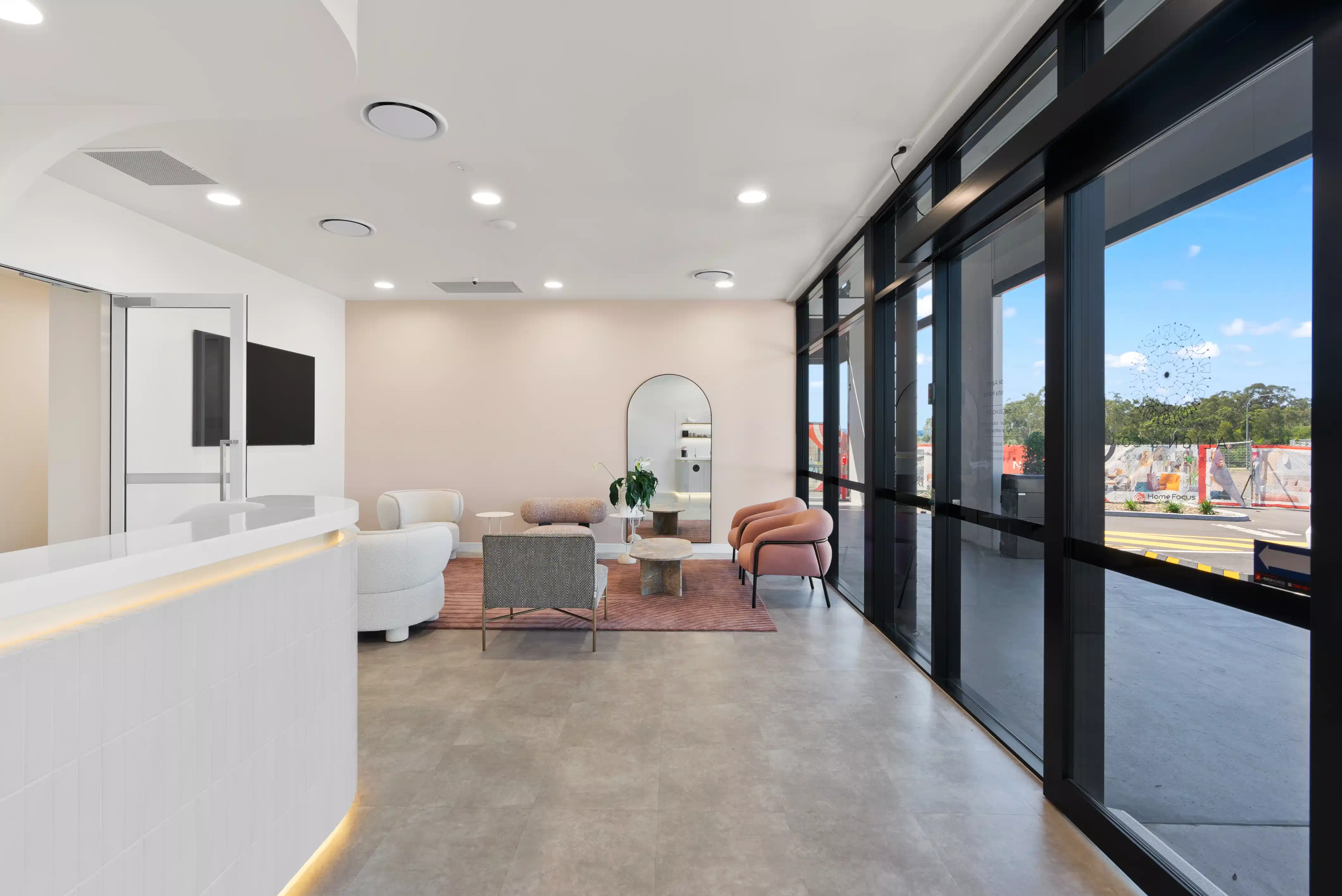 Modern waiting area with sofas, armchairs, mirror, large windows, and a white reception desk with lighting.