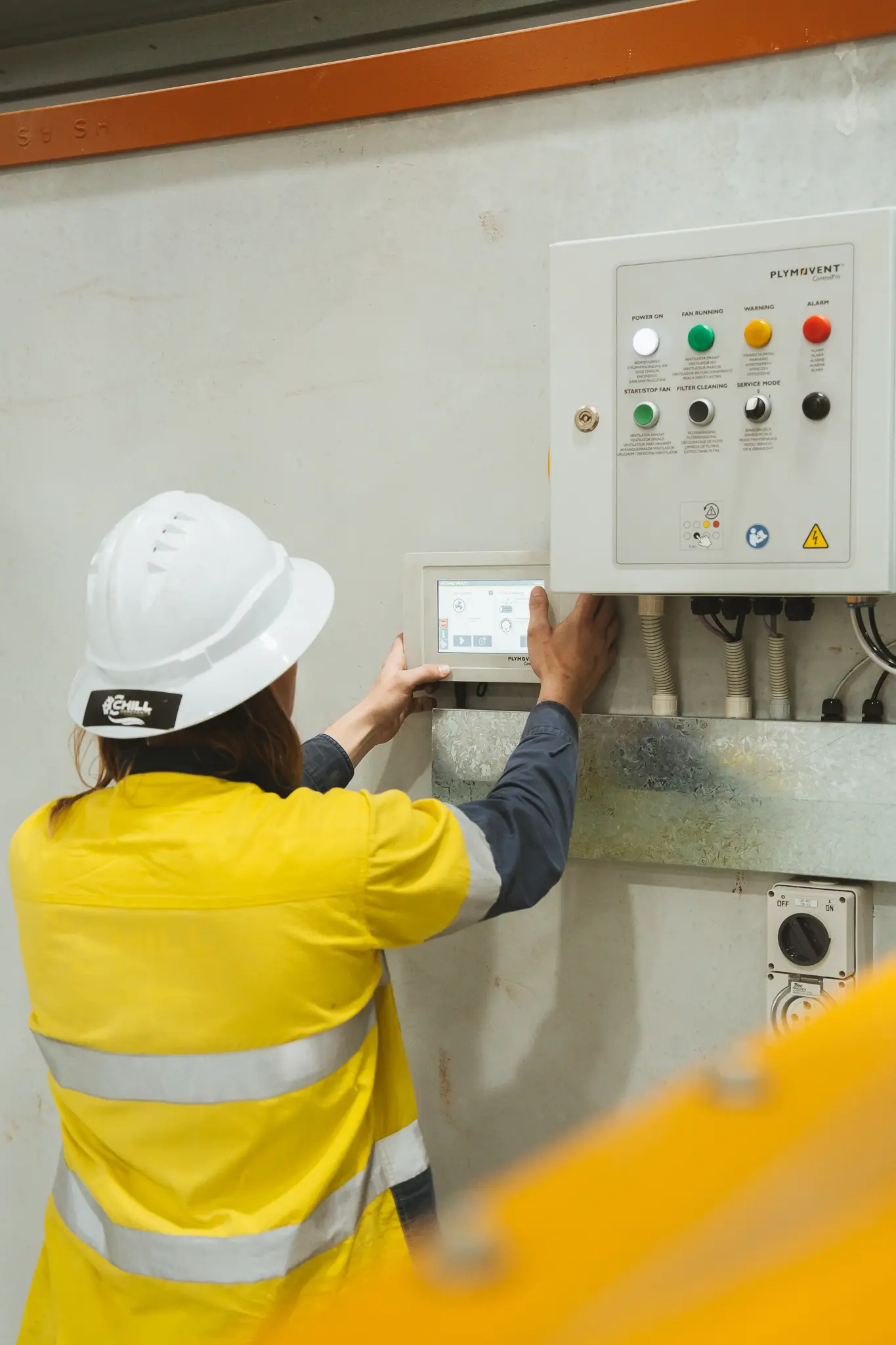 Worker in yellow reflective jacket and white hard hat adjusting industrial control panel.