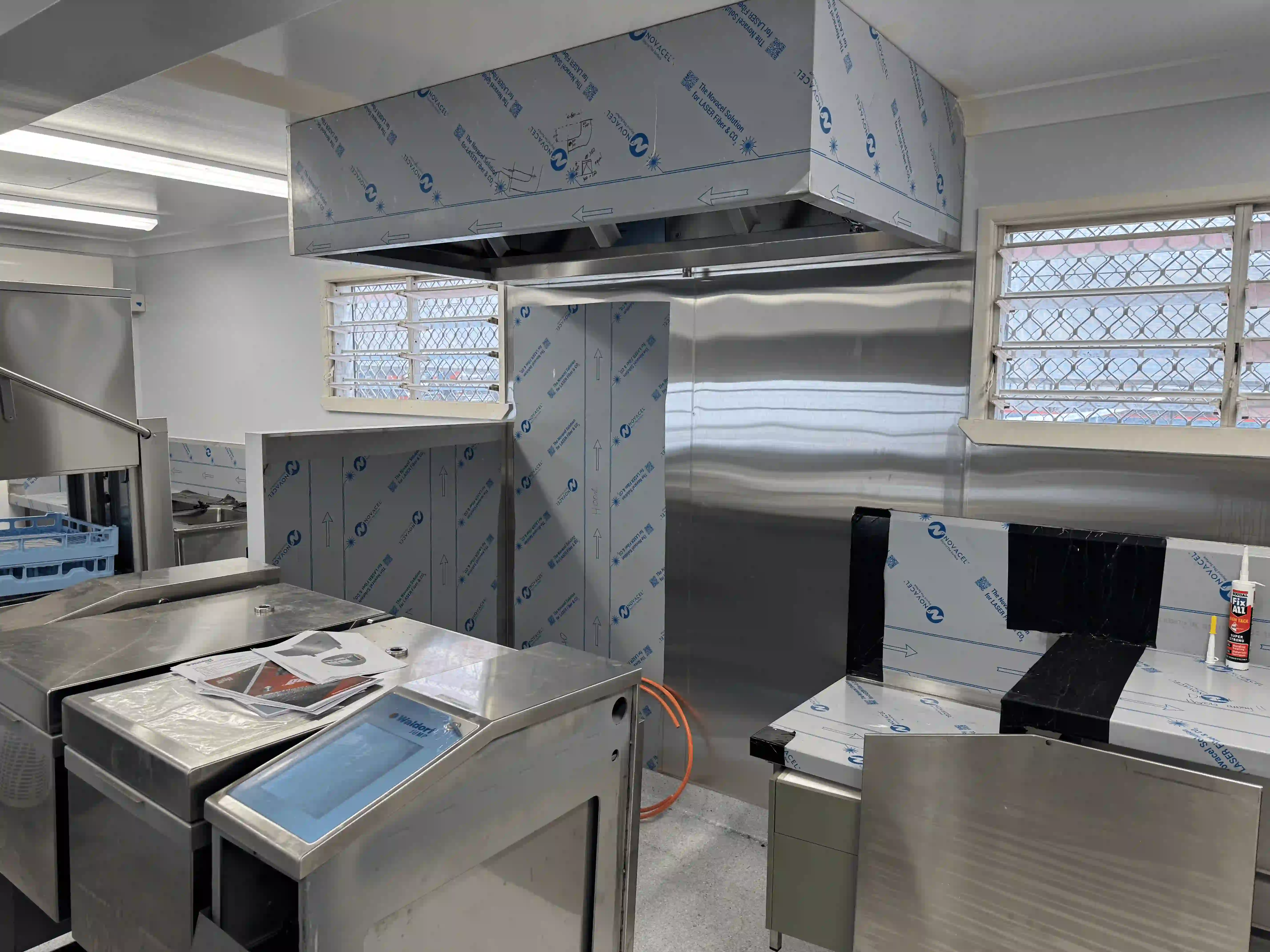 Commercial kitchen with stainless steel equipment and ventilation hood still covered in protective film.