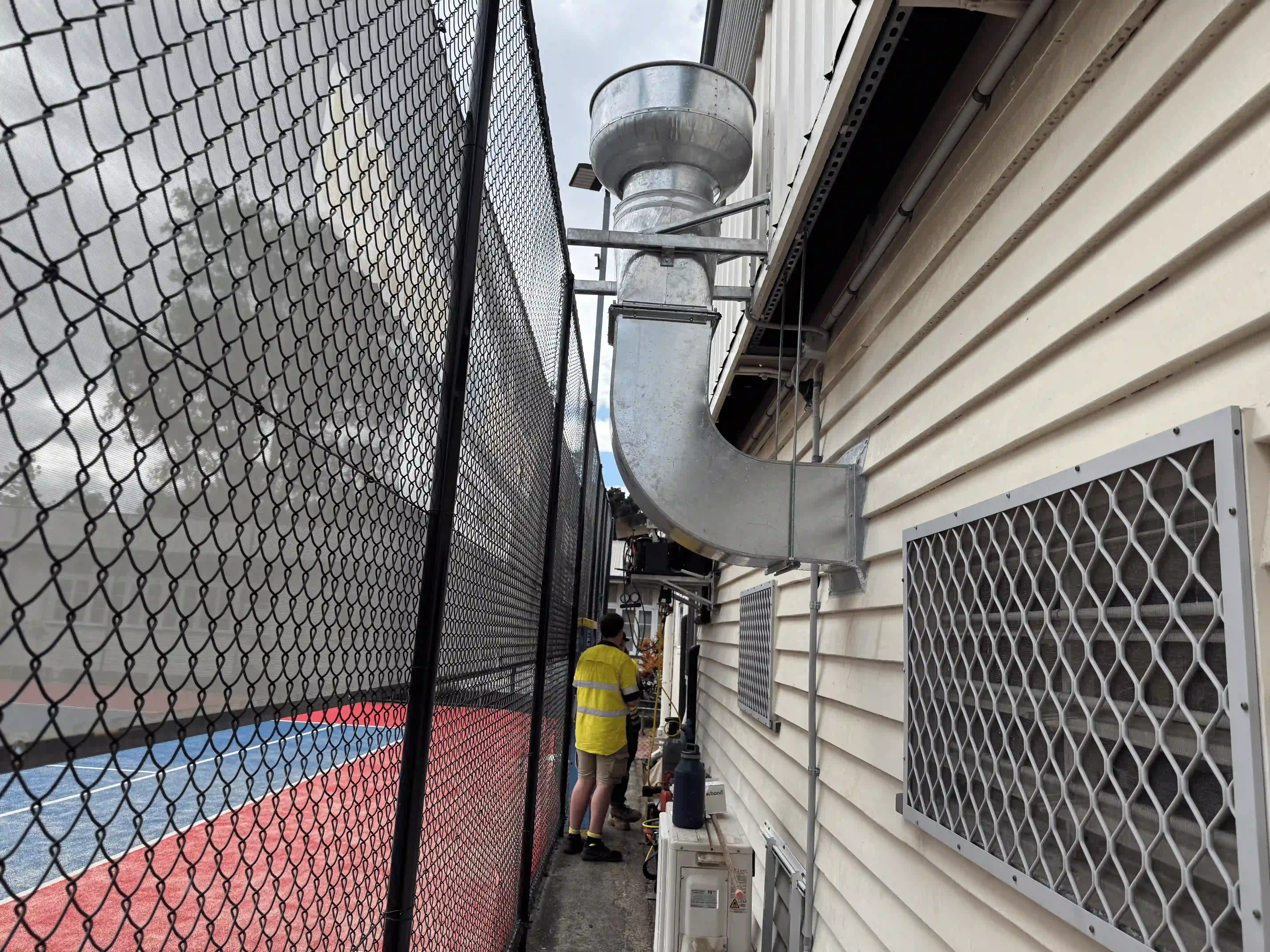Two men in yellow shirts standing between a fence and a building with ventilation ducts.