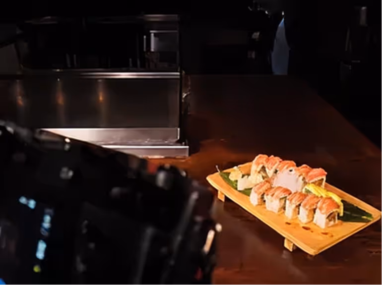 Wooden platter with sushi rolls topped with salmon on a dark table, with a blurred camera in the foreground.