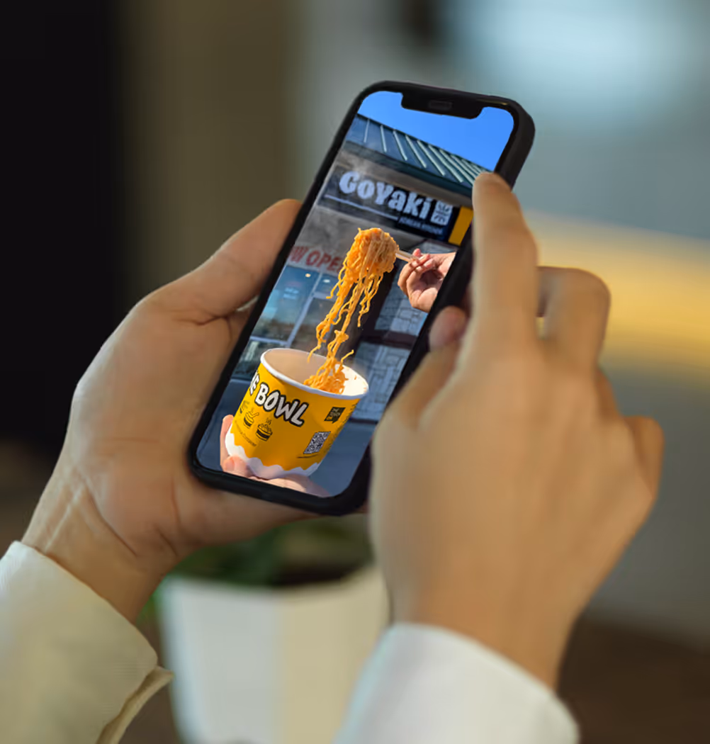 Person holding a smartphone showing a photo of chopsticks lifting noodles from a yellow bowl outside a restaurant named Goyaki.