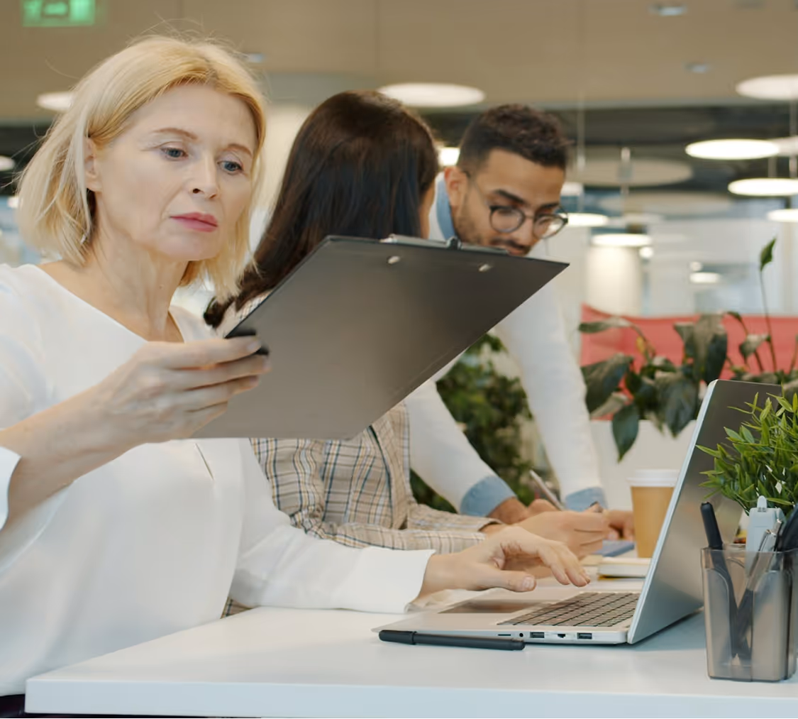 Three colleagues working together in an office; a woman in white holding a clipboard, another woman and a man writing at a desk with a laptop and plants.