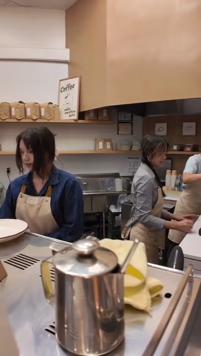 Two women wearing aprons working behind the counter in a coffee shop with bags of coffee on shelves in the background.