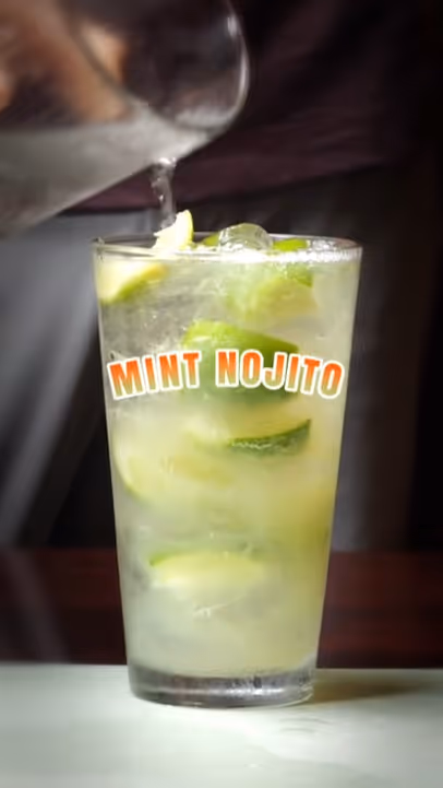 Glass of mint mojito with lime slices and ice, being topped with liquid from a pitcher.