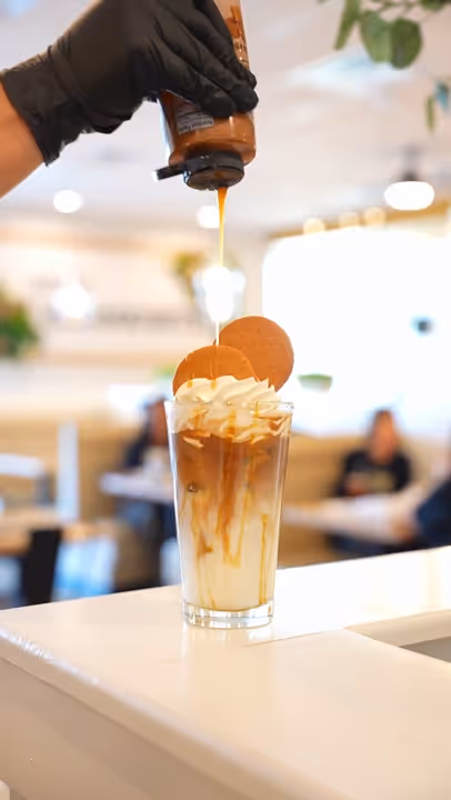 Hand wearing black glove drizzling caramel sauce onto whipped cream topped iced coffee with cookies in a glass.