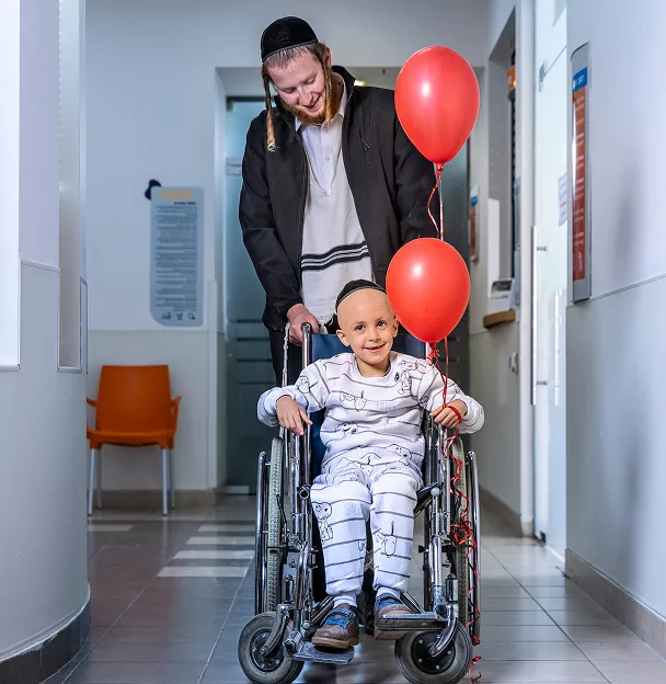 Smiling boy in pajamas sitting in wheelchair holding two red balloons, pushed by a man in traditional Jewish attire in a hospital corridor.