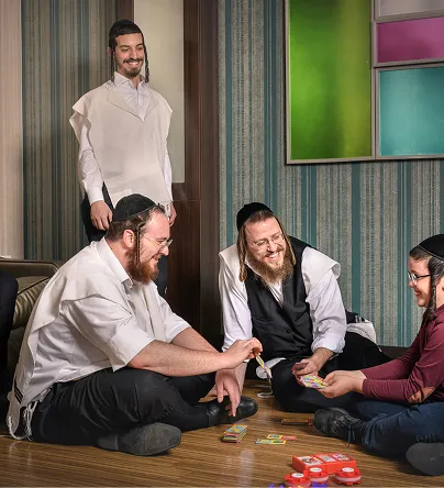 Four men, three seated on the floor playing a colorful card game and one standing, smiling in a room with striped wallpaper and framed abstract art.