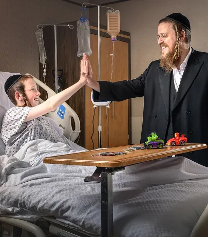 A boy in a hospital bed and a man with a beard high-five over a table with toy cars and game pieces.