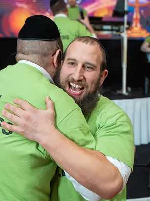 Two men wearing green shirts and black kippahs embracing in a joyful hug indoors.