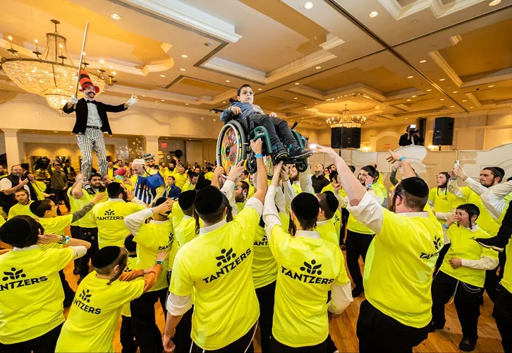 Group lifting a boy in a wheelchair wearing yellow shirts that say TANTZERS at an indoor event.