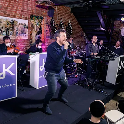 Man singing on stage with band playing instruments in a venue with brick walls and audience.