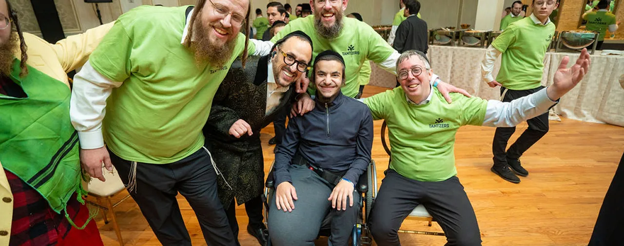 Group of smiling men, some wearing green Tantzers shirts, posing indoors, one man in wheelchair.