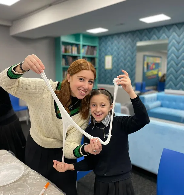 Two girls smiling and stretching slime together in a cozy room with bookshelves and blue sofas.