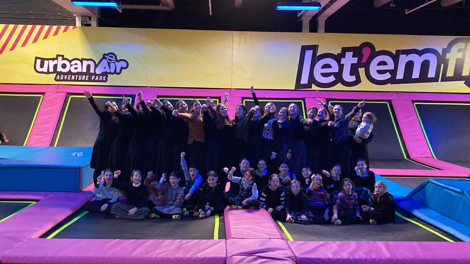 Group of adults and children posing and raising arms at Urban Air Adventure Park trampoline area with bright pink and blue padding and colorful signage in the background.