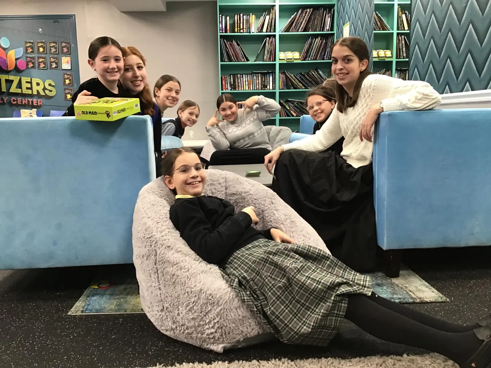 Group of eight smiling girls and young women in a cozy room with blue couches and bookshelves, one girl reclining on a plush bean bag in front.