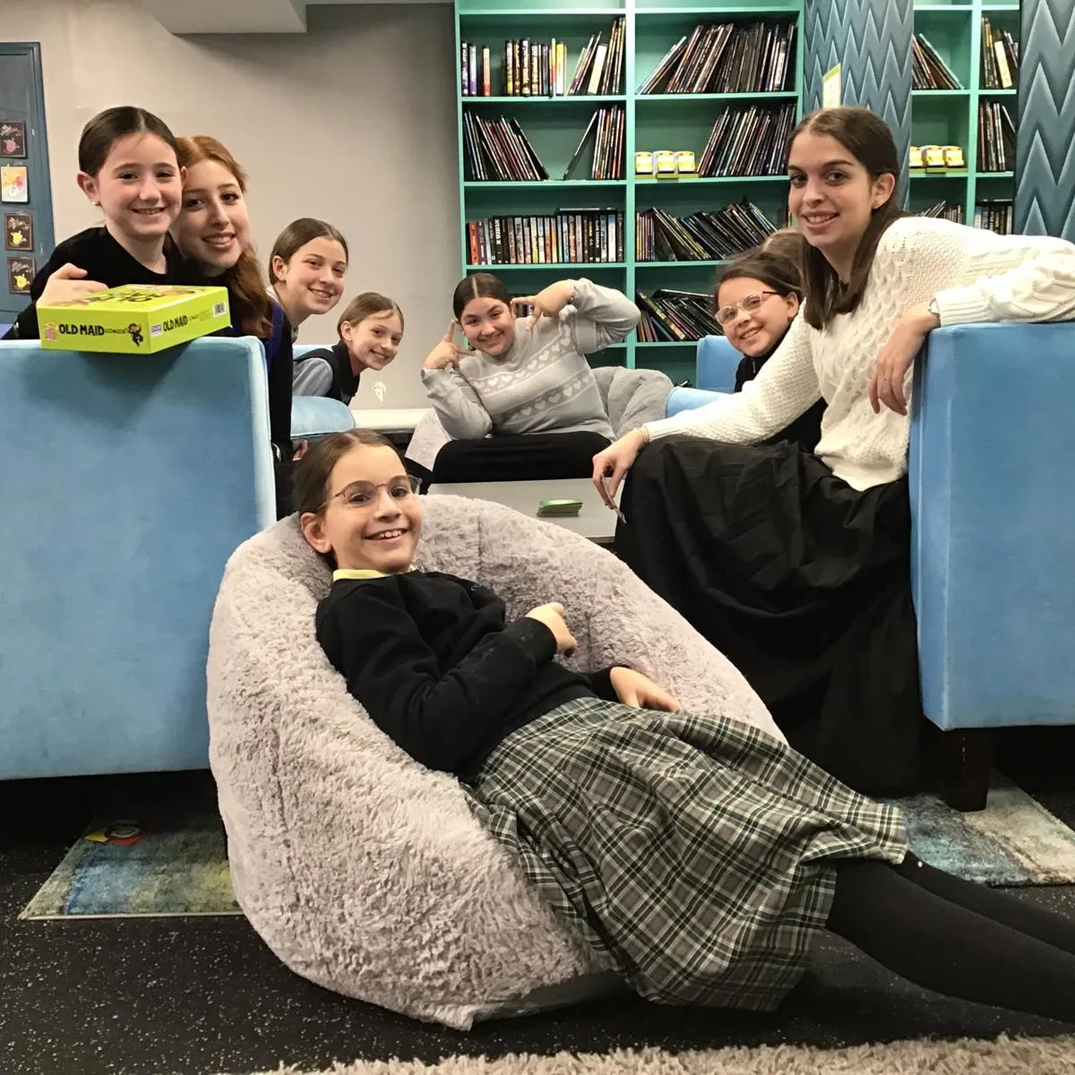 Group of eight smiling girls sitting and lounging in a cozy room with blue couches and bookshelves filled with books, one girl lying comfortably on a plush gray bean bag chair in the foreground.